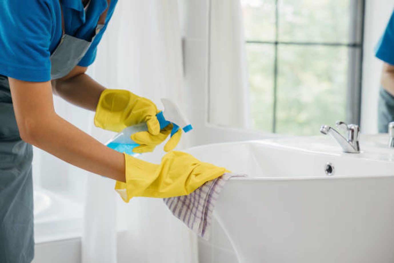 A maid wearing gloves diligently cleans bathroom sink and faucet eliminating limescale with spray detergent. Her focus on housework ensures purity hygiene and shining fixtures. spray cleaner